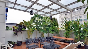 Un área de estar al aire libre cubierta, con plantas en macetas, sillas de metal y un banco.