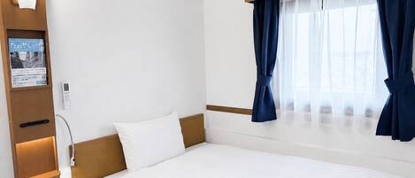 A hotel room with a bed, a bedside table, a wall-mounted light, a window with curtains, and a wall-mounted TV.