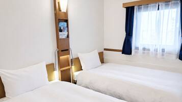 Una habitación de hotel con dos camas, una mesita de noche, estante empotrado con material de lectura y ventana con cortinas.