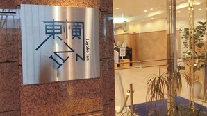 Entrada de un hotel con un letrero en japonés e inglés, mostrador de recepción y plantas en macetas.