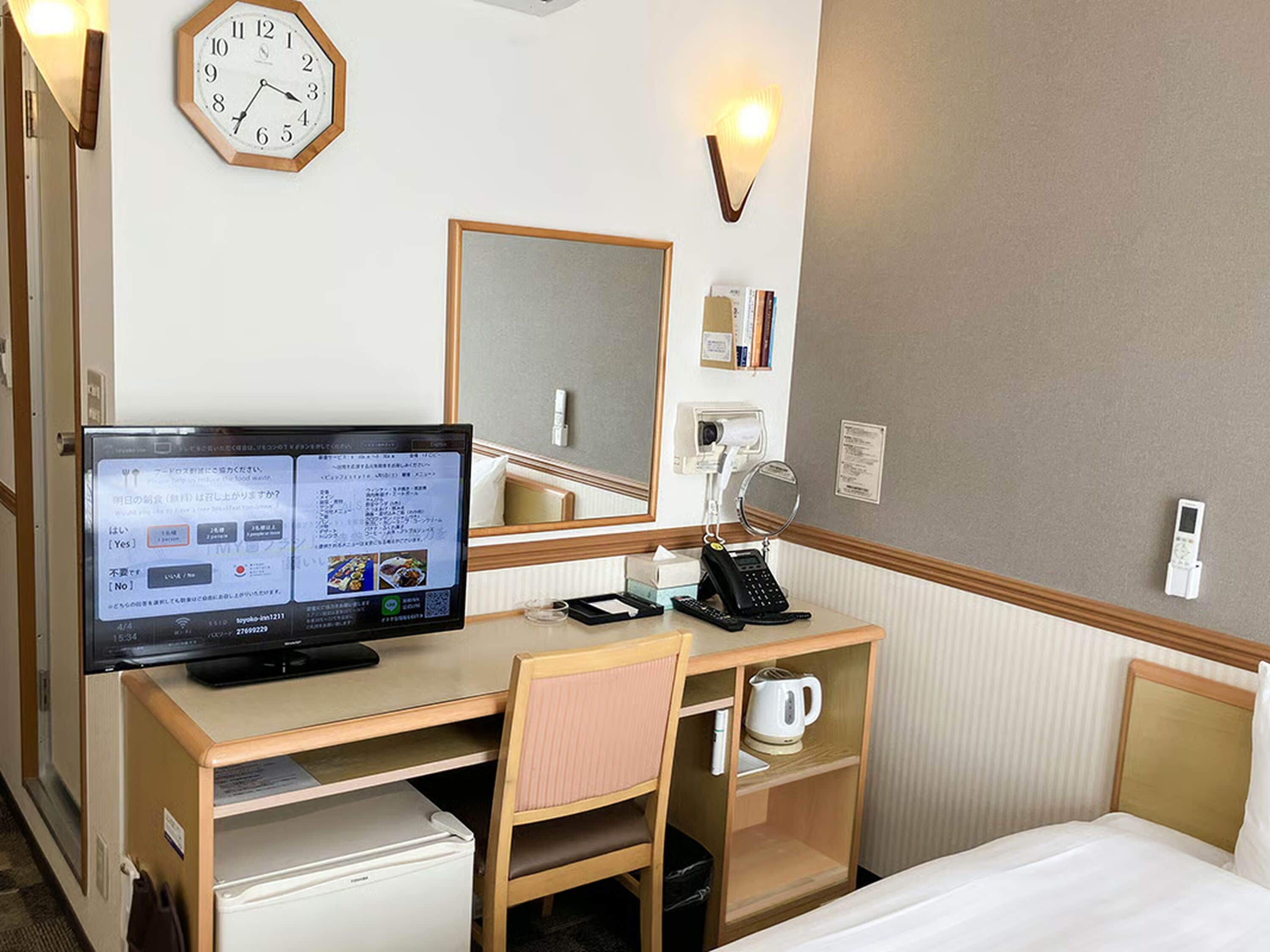 A hotel room with a desk, chair, bed, clock, and various amenities including a kettle, phone, and a mirror.