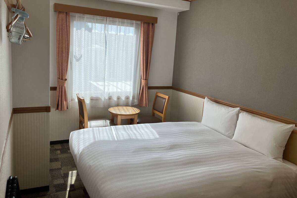 Ett hotellrum med en säng, en stol, ett litet bord och ett fönster med gardiner.