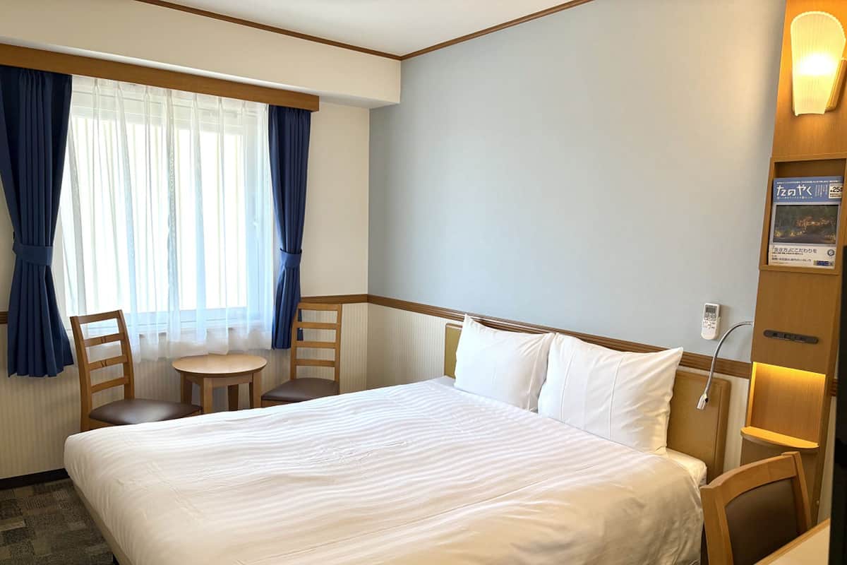 A hotel room with a large bed, a small table, two chairs, a window with curtains, and a wall-mounted light.