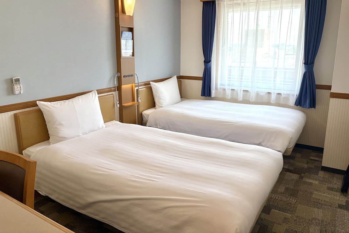 A hotel room with two beds, a bedside table, a lamp, a telephone, and a window with curtains.