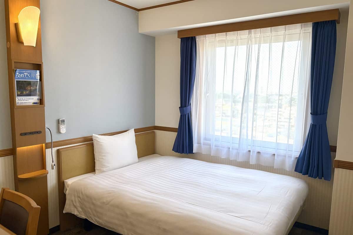 A hotel room with a large bed, a bedside table, a lamp, a window with curtains, and a view of the outdoors.