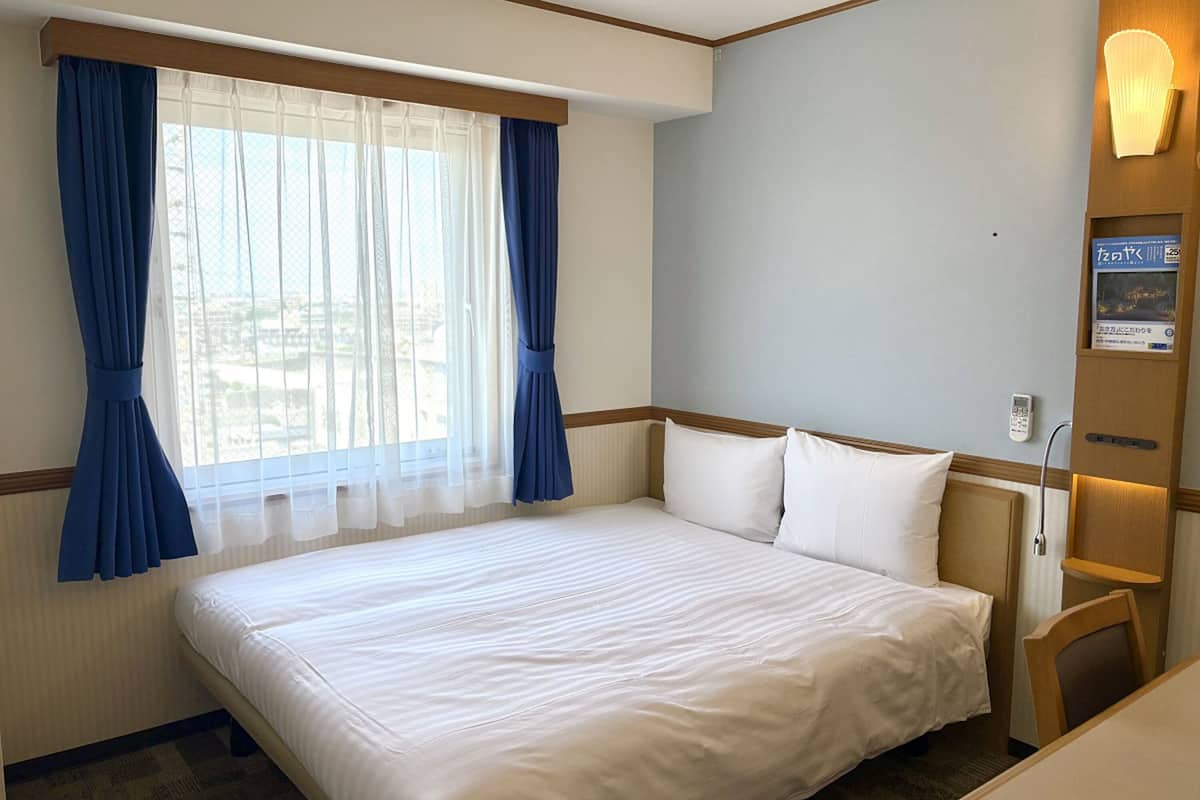A hotel room with a large bed, a window with blue curtains, a bedside table, and a wall-mounted light.