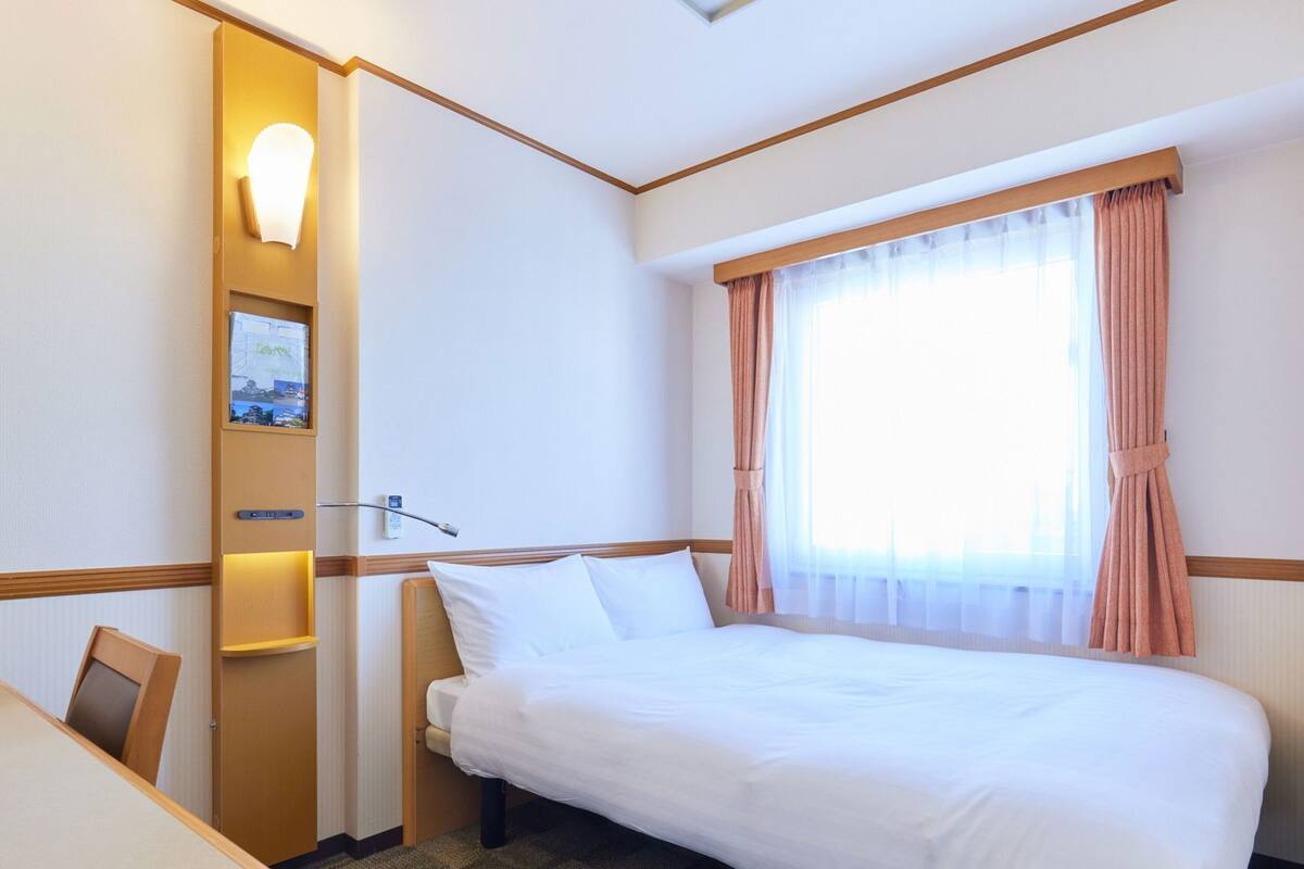 A hotel room with a bed, a desk, a chair, a wall-mounted TV, and a window with curtains.