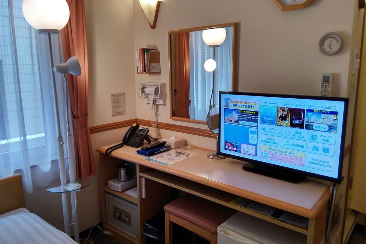 A hotel room with a desk, a computer, a telephone, a clock, and a mirror.