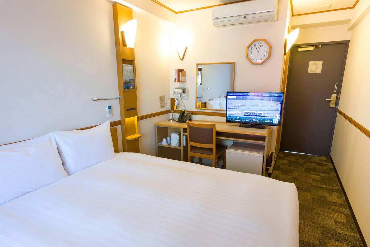 A hotel room with a bed, desk, chair, computer, and a clock on the wall.