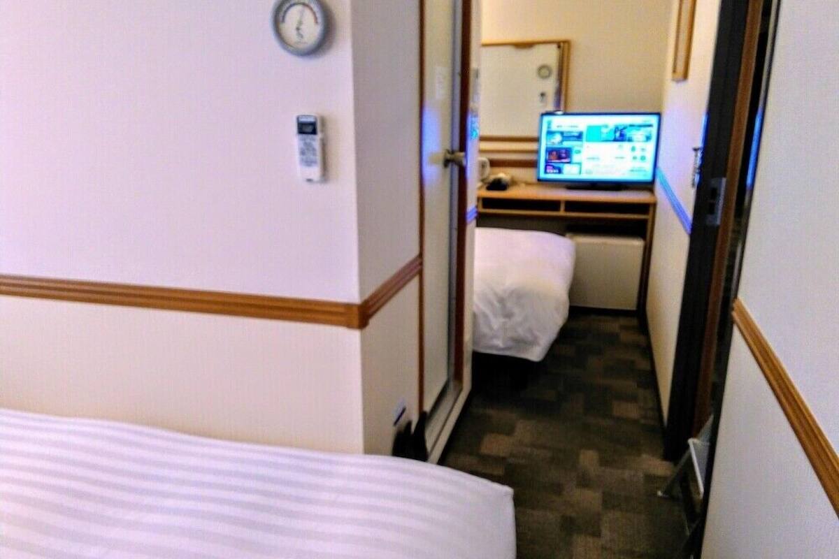 A hotel room with a bed, a desk with a computer, a clock, and a door.