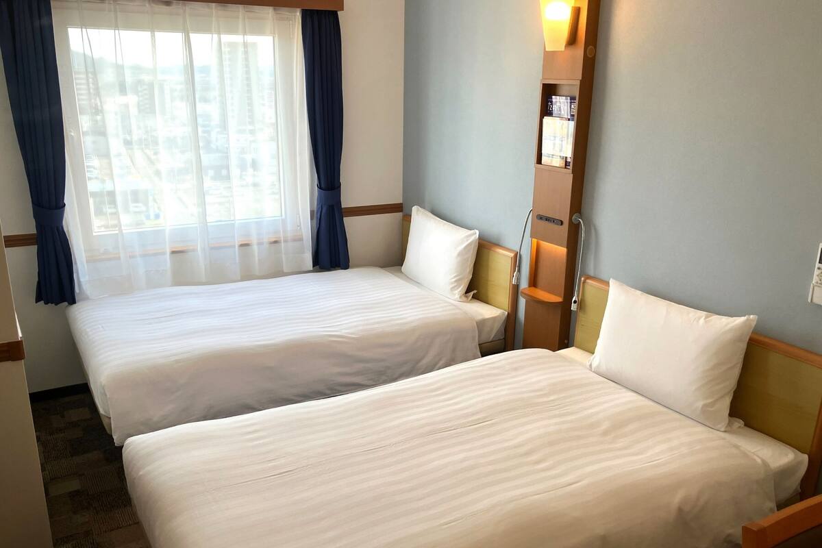 Ein Hotelzimmer mit zwei Einzelbetten, einem Fenster mit durchsichtigen Vorhängen, einem an der Wand befestigten Regal mit Lektüre und einem Nachttisch mit Lampe.