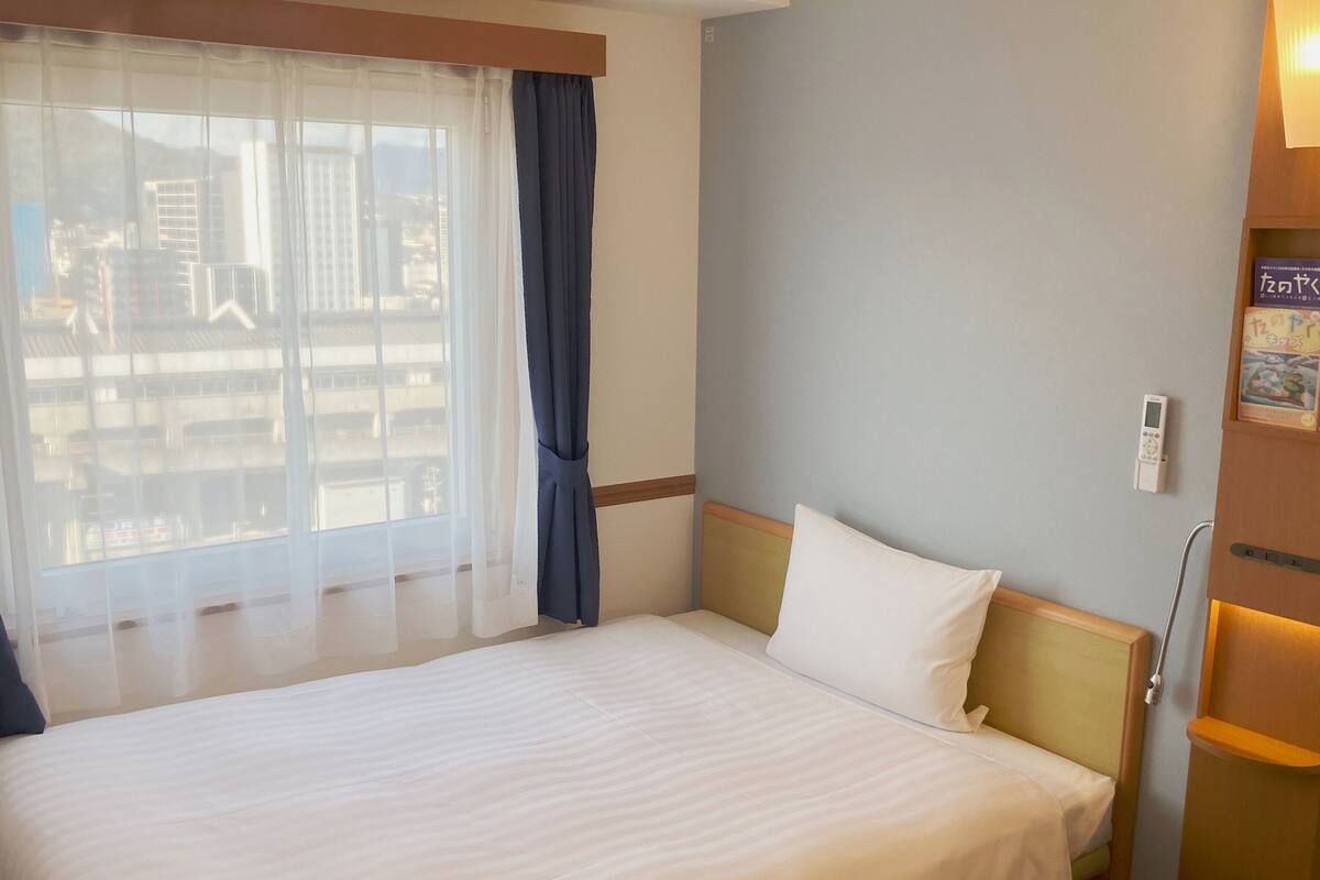 Ein Hotelzimmer mit einem großen Bett, einem Fenster mit durchscheinenden Vorhängen und einem Nachttisch mit einer Lampe.