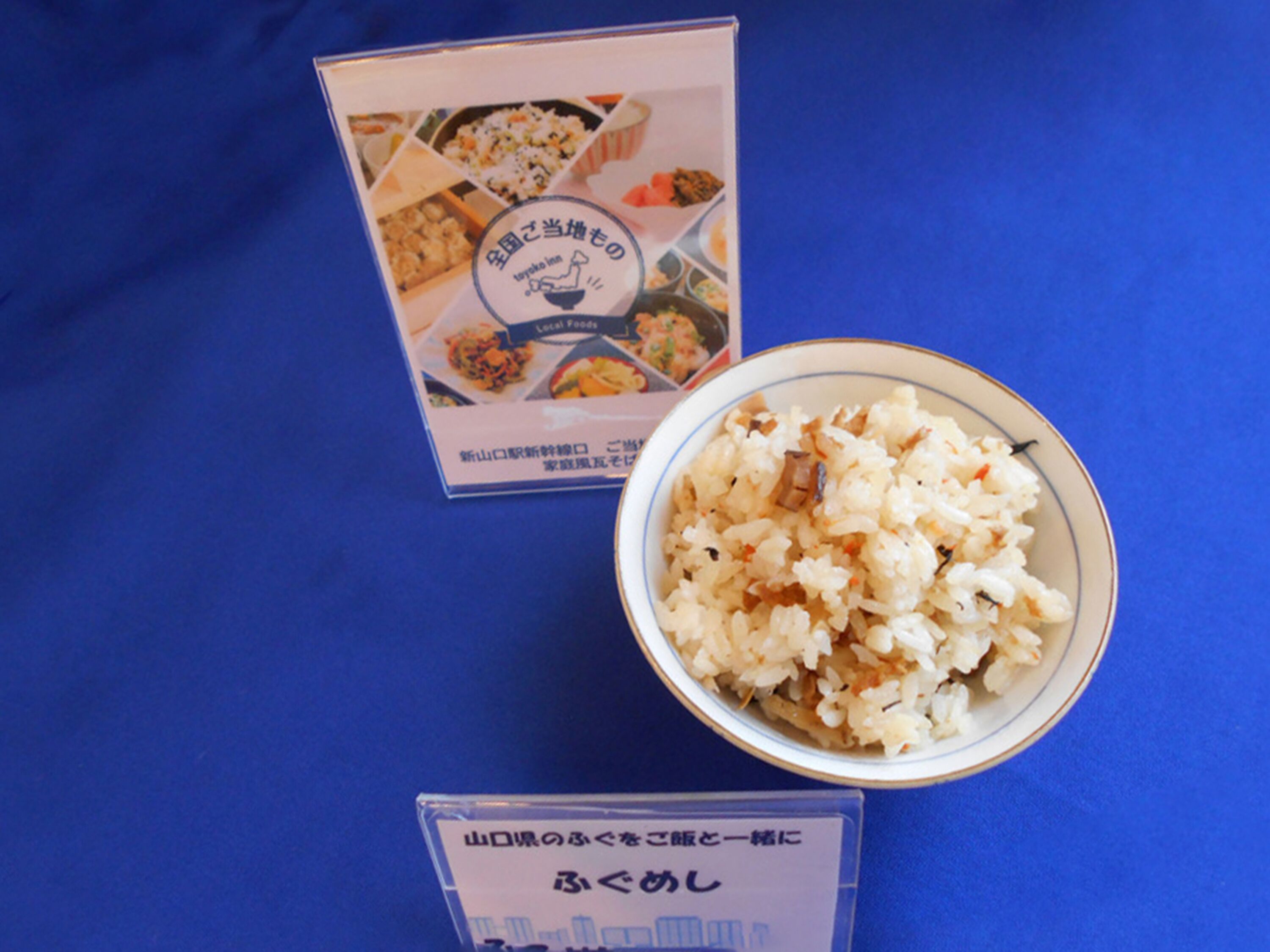 Un tazón de arroz con vegetales sobre un fondo azul, un menú con fotos de comida y una tarjeta pequeña con texto.