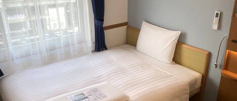 A hotel room with a bed, a window with curtains, a wall-mounted thermostat, and a wooden cabinet.