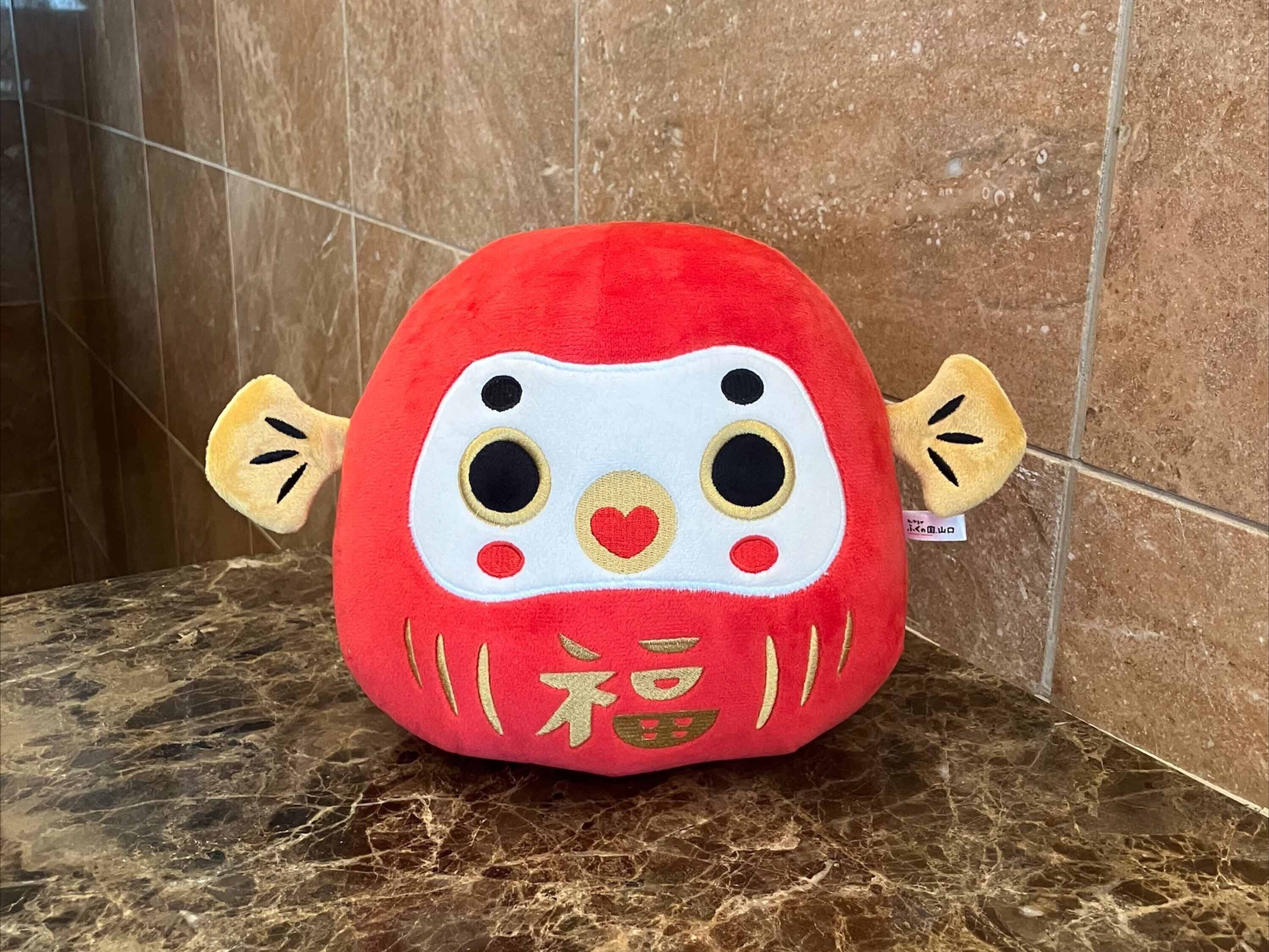 Un jouet en peluche avec un corps rouge, un visage blanc et des détails dorés, posé sur une surface en marbre devant un mur carrelé.