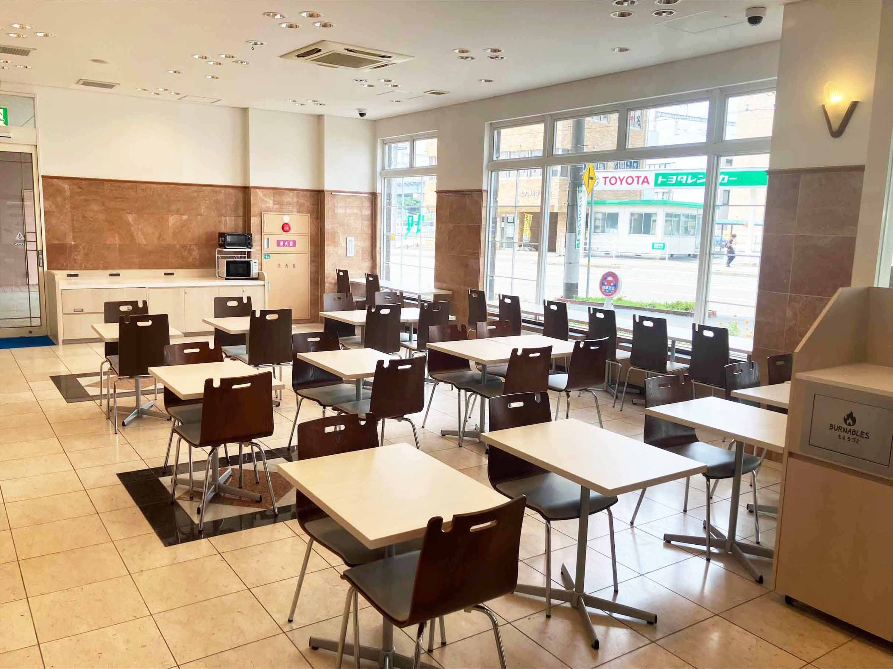 A restaurant with tables and chairs, a kitchen area, and large windows with a view of the street.