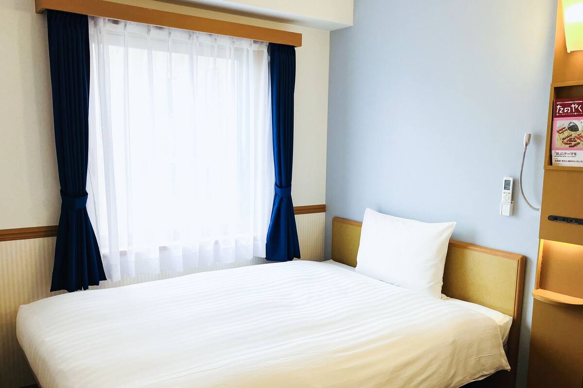 Una habitación de hotel con una cama grande, una mesita de noche, cortinas en una ventana y un termostato en la pared.