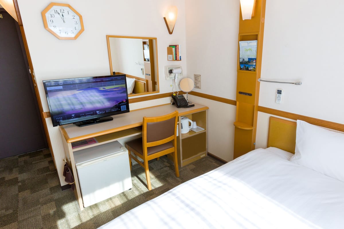 A hotel room with a bed, a desk with a computer, a chair, a small refrigerator, a clock, and a mirror.