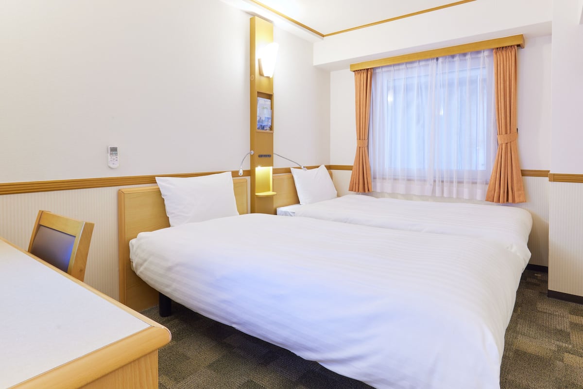 A hotel room with two beds, a desk, a bedside table, a lamp, a window with curtains, and a wall-mounted phone.