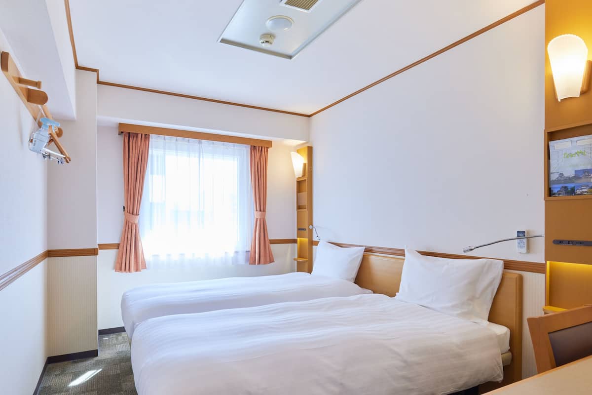 A hotel room with two beds, a window with curtains, a bedside table, and a wall-mounted lamp.
