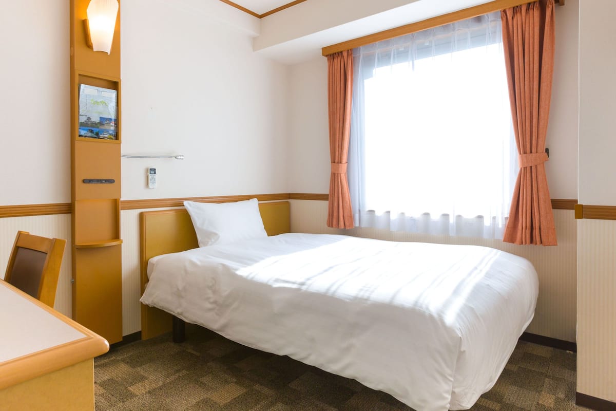 A hotel room with a bed, a desk, a chair, a wall-mounted shelf with a lamp, and a window with curtains.