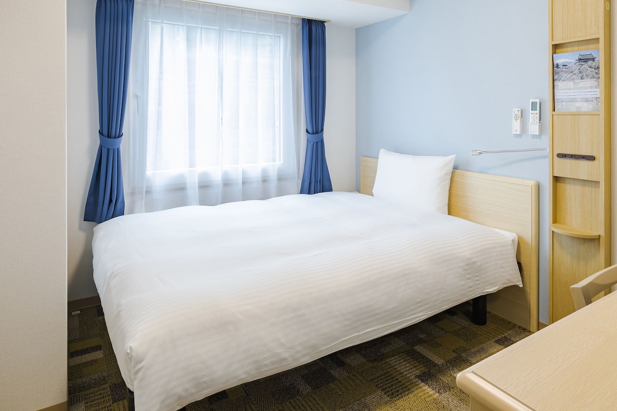 Una habitación de hotel con una cama grande, una mesita de noche, un armario de madera y una ventana con cortinas azules.