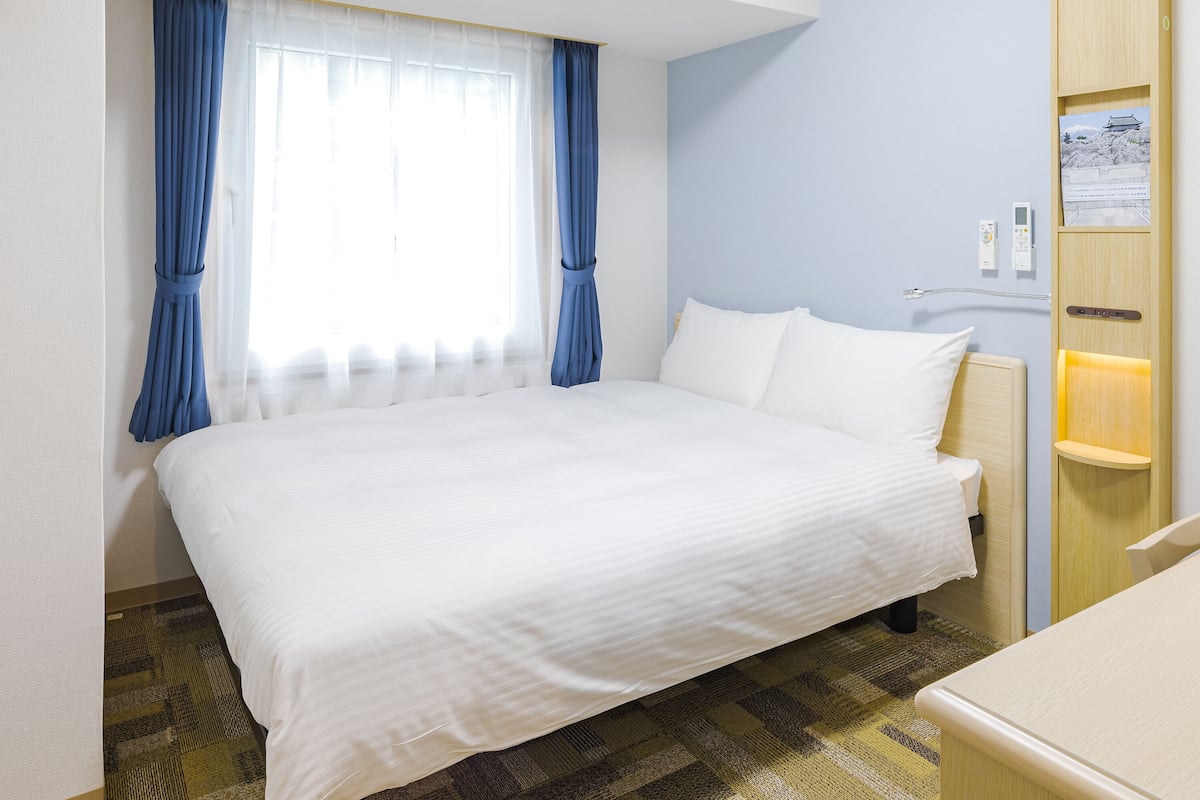 Una habitación de hotel con cama, mesita de noche, cortinas en la ventana y panel de control en la pared.