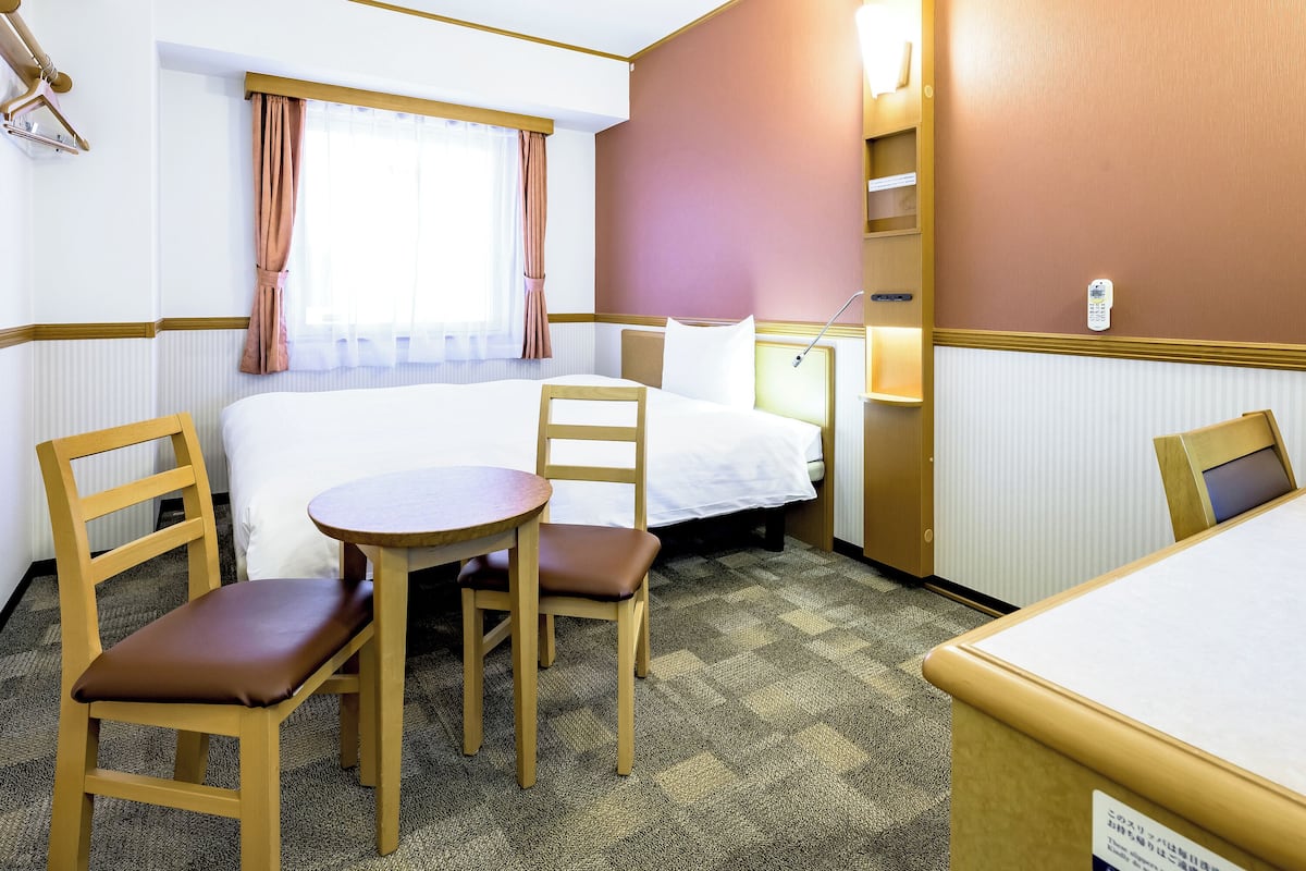A hotel room with a bed, two chairs, a small table, a window with curtains, a bedside lamp, and a wall-mounted phone.