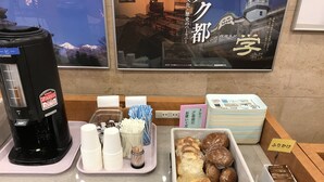 A coffee station with a coffee maker, disposable cups, and a tray with utensils. There are also stacks of disposable plates and a basket of pastries.