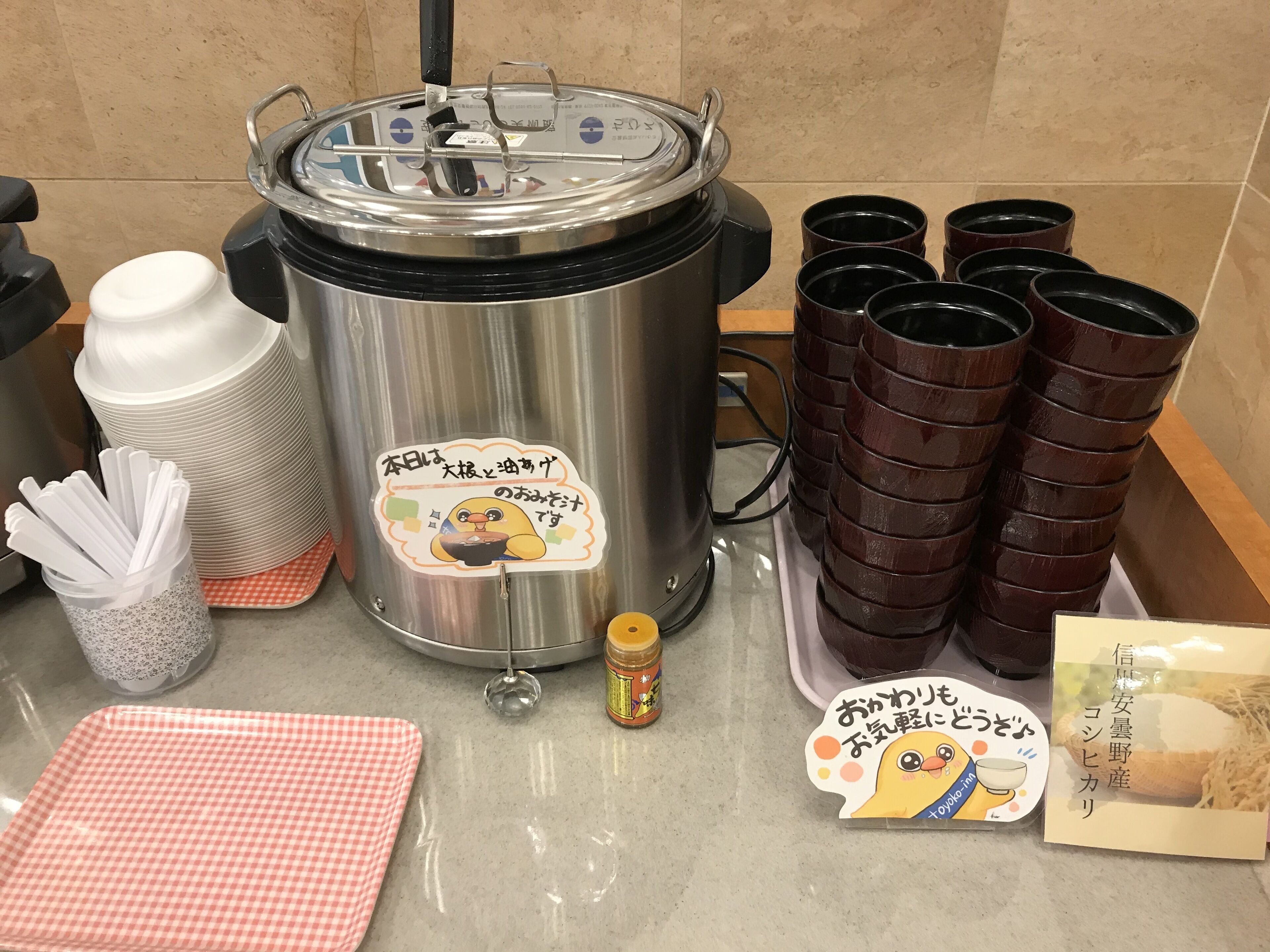 A kitchen setup with a rice cooker, stacked cups, a jar of condiments, a checkered cloth, and a menu.
