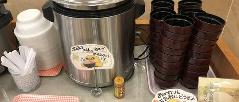A kitchen setup with a rice cooker, stacked cups, a jar of condiments, a checkered cloth, and a menu.