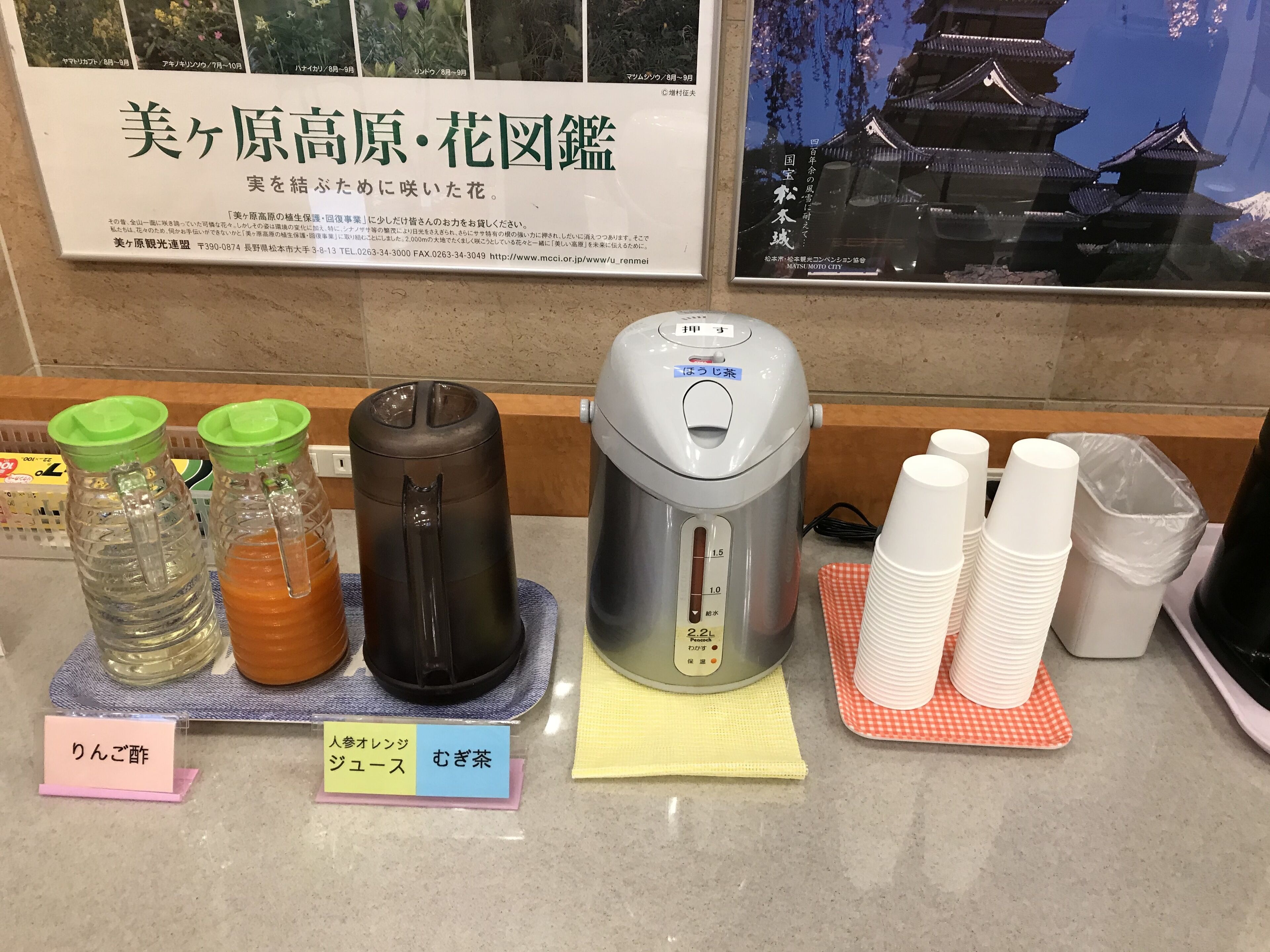 A kitchen counter with a rice cooker, two thermoses, and stacked paper cups.