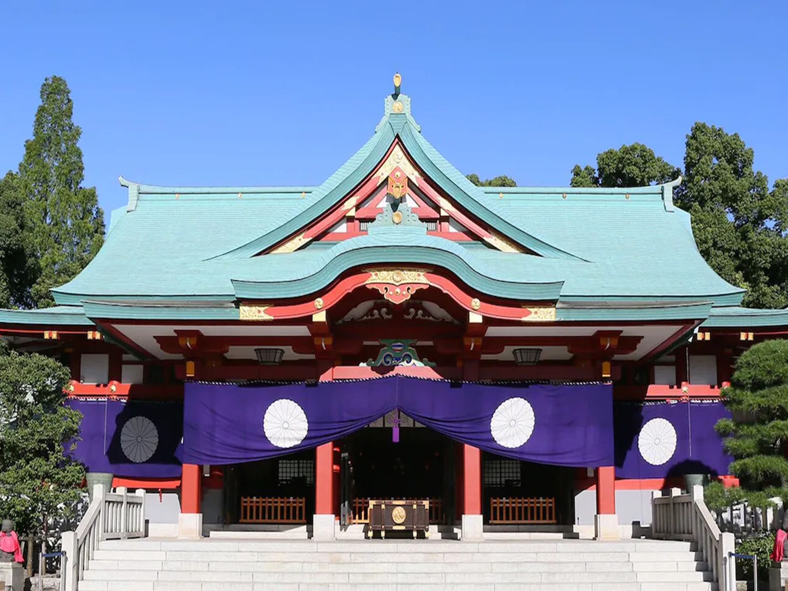 Un bâtiment traditionnel japonais doté d’un toit végétalisé, de murs rouges et blancs et d’un auvent d’entrée violet.