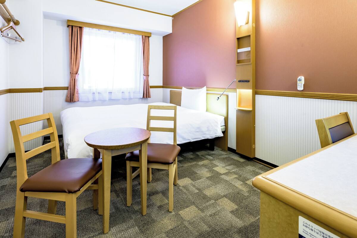 A hotel room with a bed, two chairs, a small table, a window with curtains, a bedside lamp, and a wall-mounted phone.