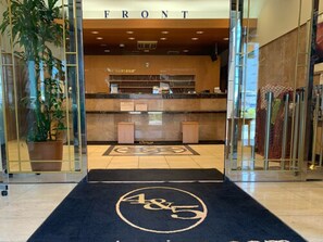 Un espace d’accueil d’hôtel comprenant un comptoir de réception, une plante en pot et un tapis orné d’un logo.