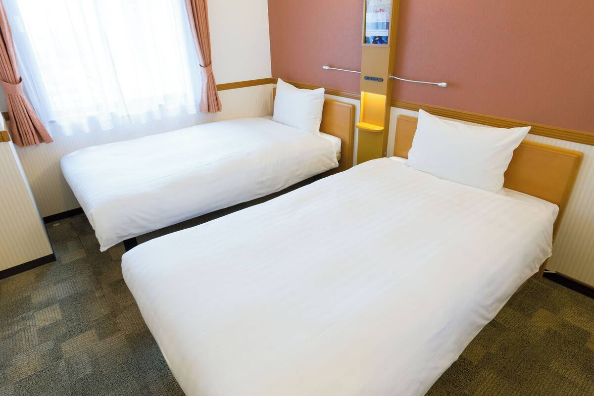 A hotel room with two single beds, a small bedside table, a nightstand, a window with curtains, and a wall-mounted light.