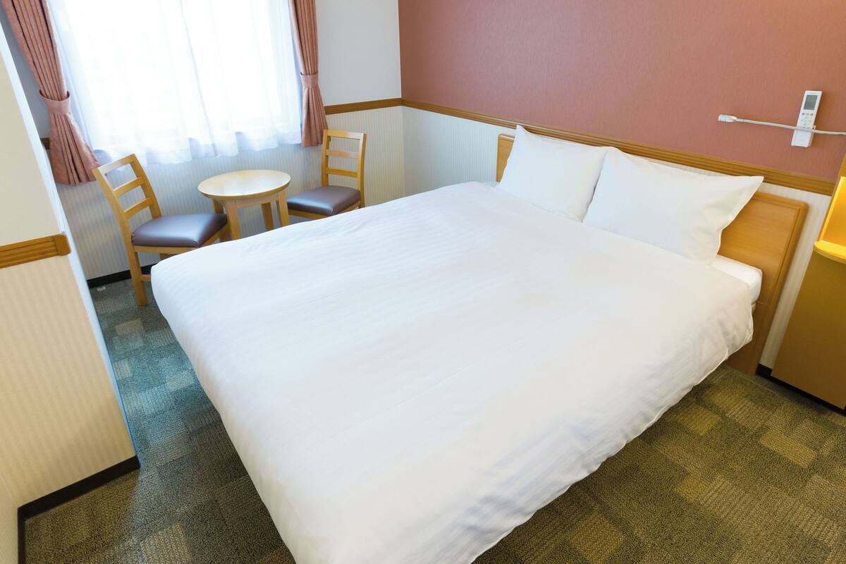 A hotel room with a large bed, a small round table, two chairs, and a bedside table with a lamp.
