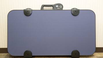 Une valise bleue avec des poignées et des roulettes noires, posée sur un sol recouvert d’une moquette, appuyée contre un mur.
