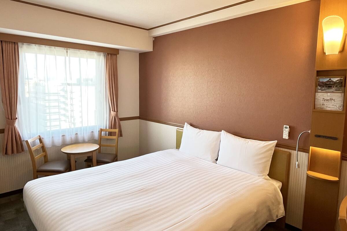 A hotel room with a large bed, a small table, two chairs, a window with curtains, and a wall-mounted light fixture.