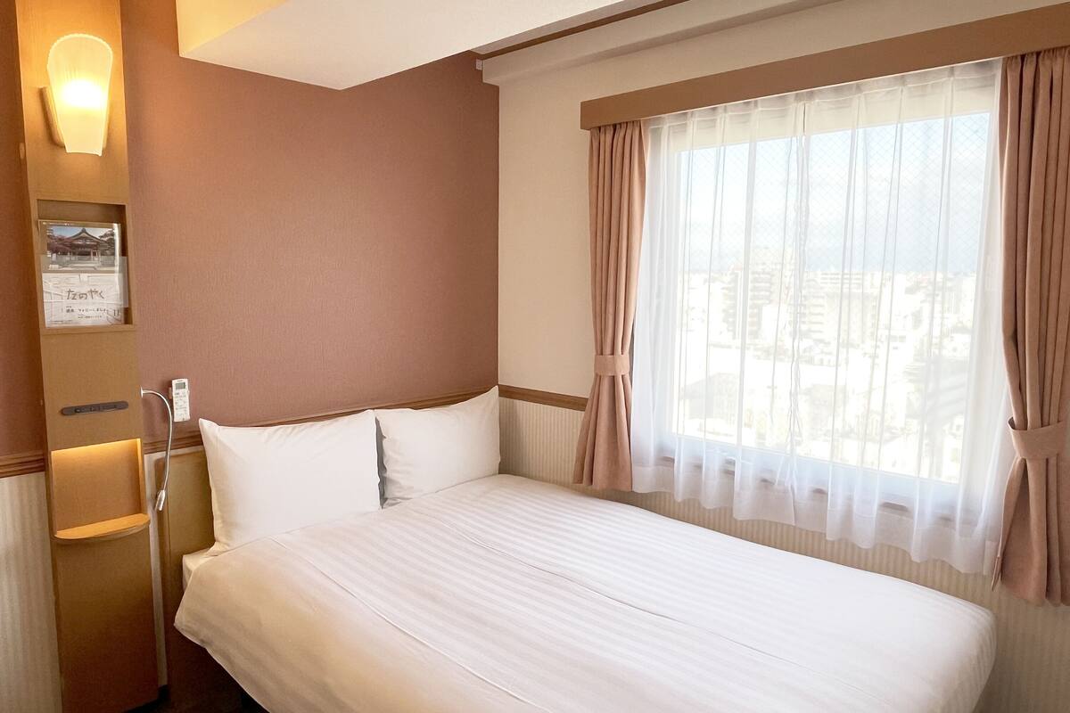 A hotel room with a large bed, a window with sheer curtains, a wall-mounted light, and a small shelf with a framed picture.