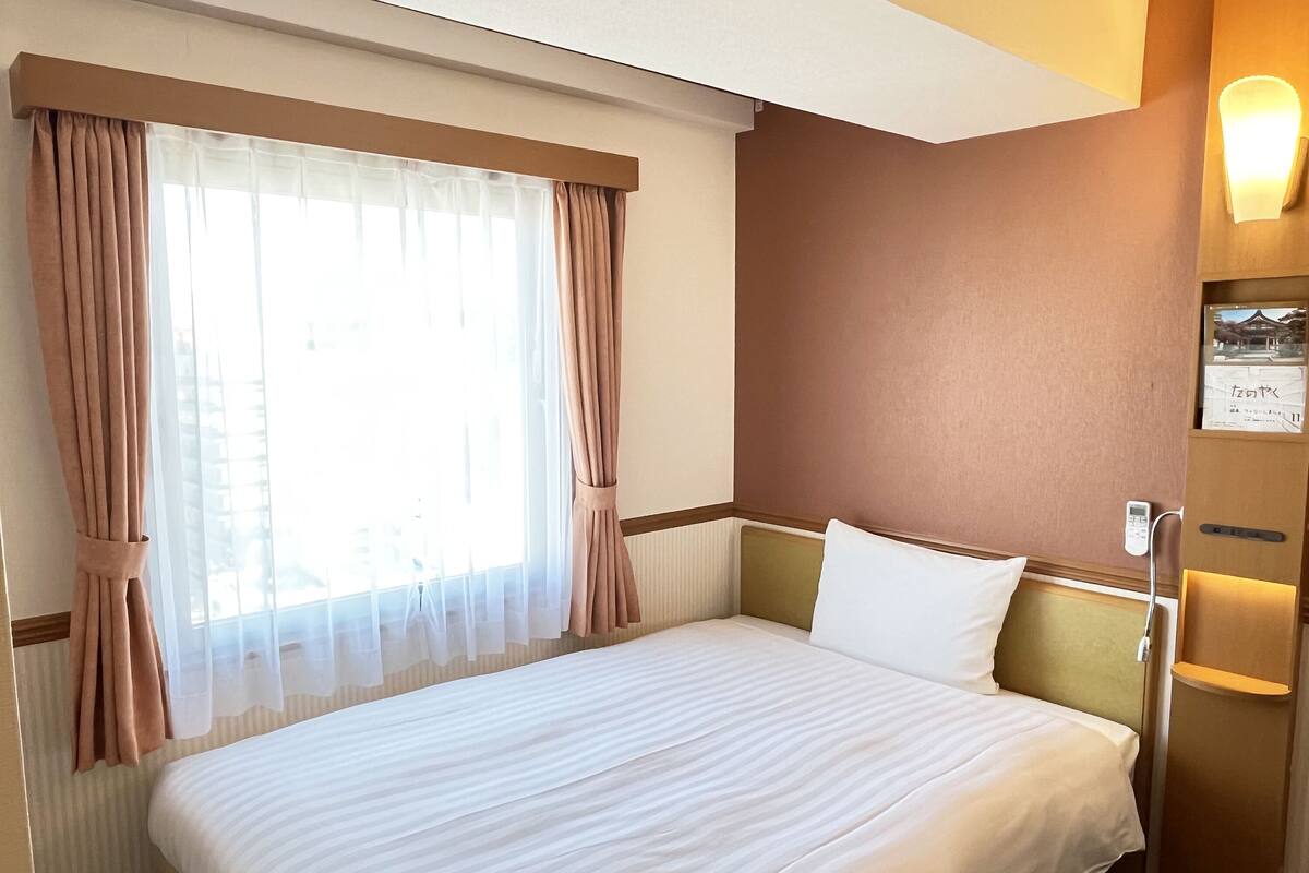 A hotel room with a bed, curtains, a window, a wall-mounted light, and a small shelf with a picture.