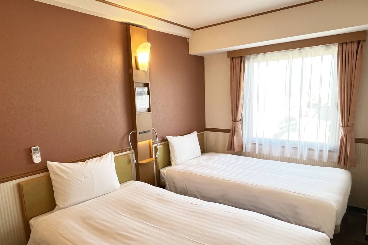 A hotel room with two beds, a bedside table, a lamp, and a window with curtains.