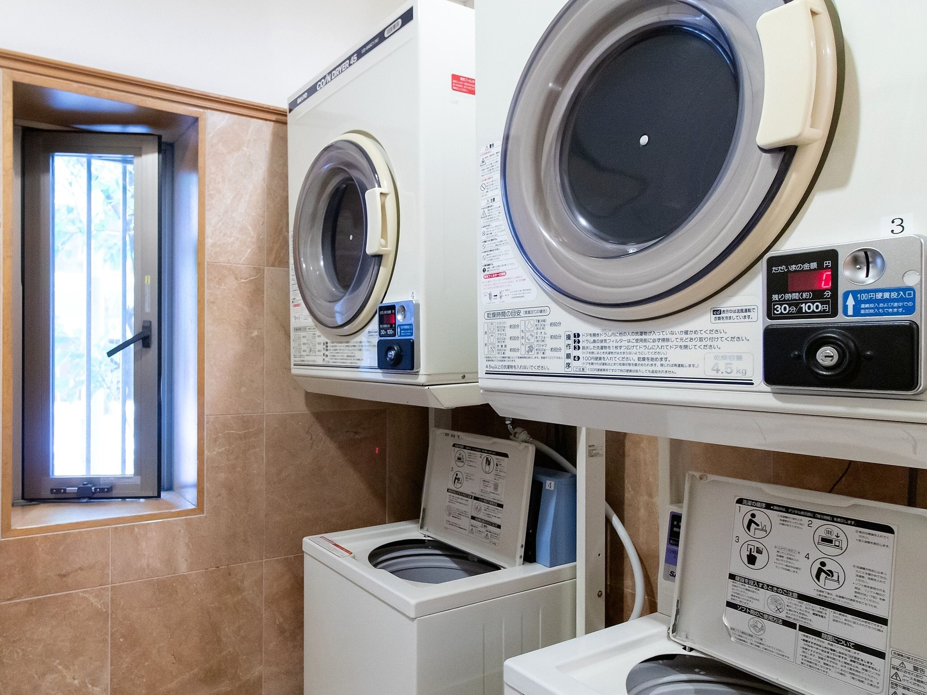 A laundry room with two front-loading washing machines, a dryer, and a window with a view of trees.