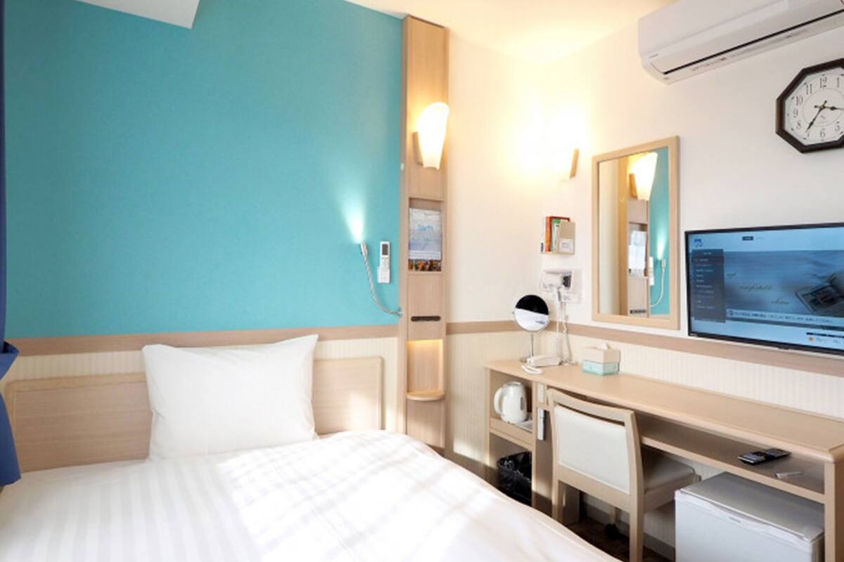 A hotel room with a bed, a desk, a mirror, a television, and an air conditioning unit.