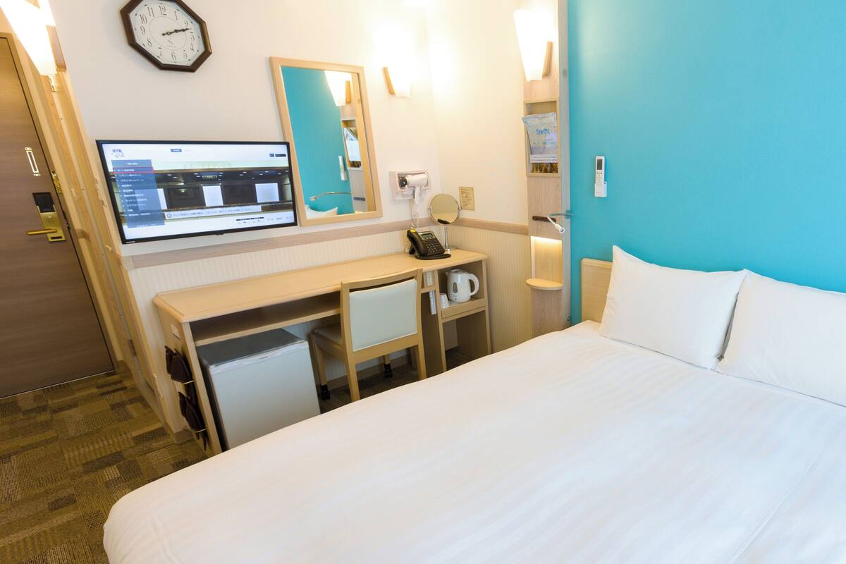 A hotel room with a bed, a desk, a computer, a clock, and a mirror.