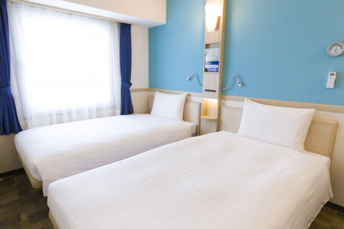 A hotel room with two beds, a window with blue curtains, a wall clock, and a bedside table with a lamp.