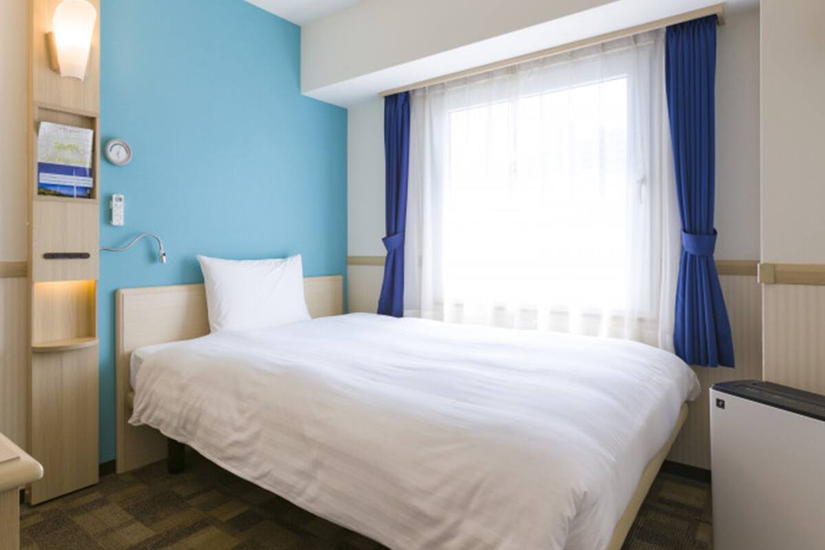 A hotel room with a bed, a nightstand, a lamp, a window with blue curtains, and a wall-mounted clock.