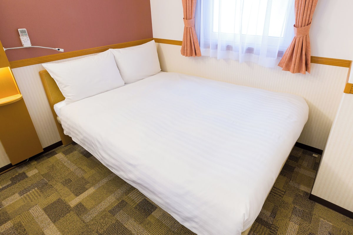 A hotel room with a neatly made bed, a bedside lamp, a telephone, and a window with curtains.