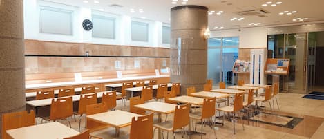 A spacious cafeteria with multiple tables and chairs, a clock on the wall, and a pillar in the center.