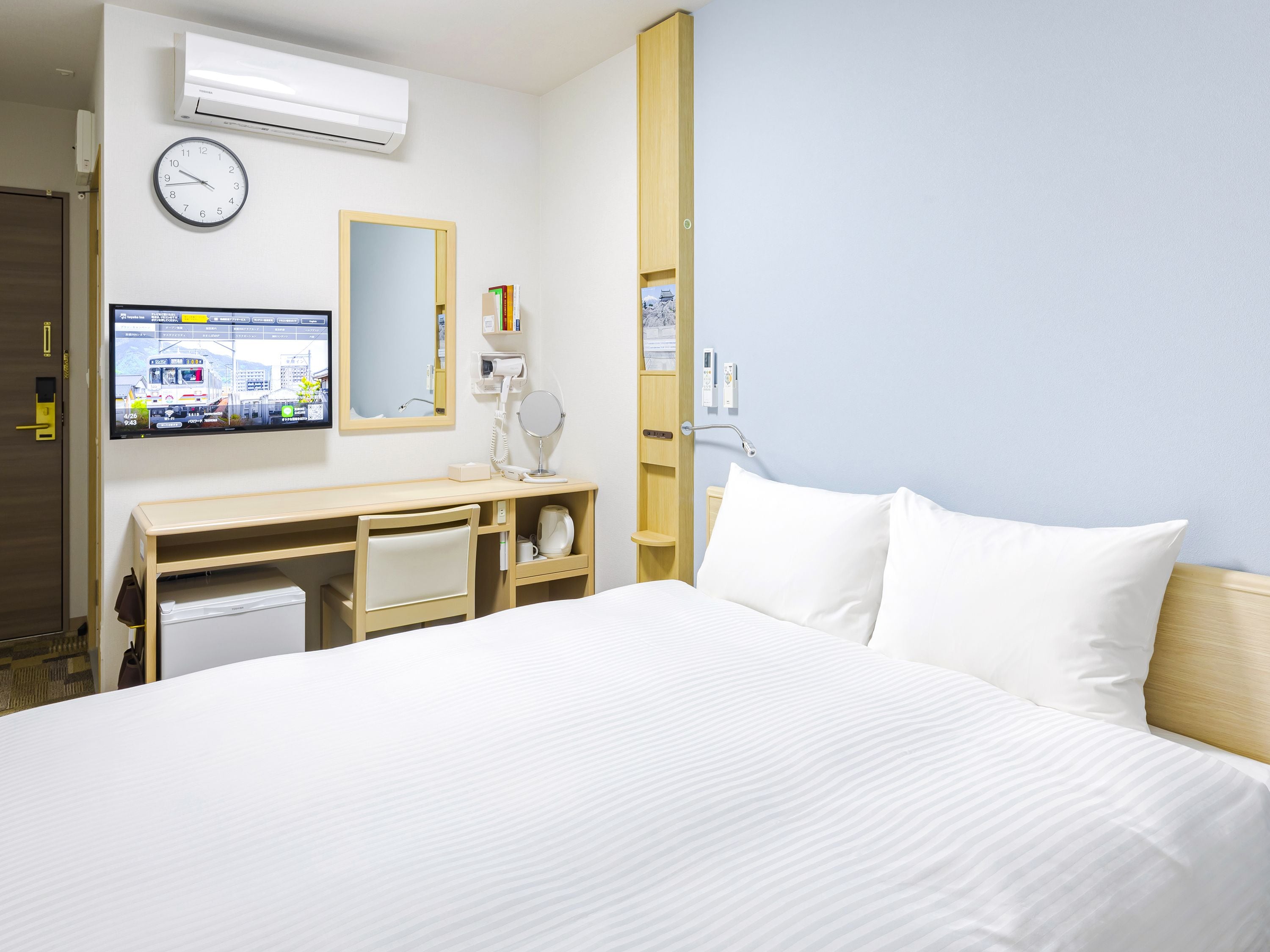 Habitación de hotel con cama, escritorio con monitor de ordenador, espejo y aire acondicionado.